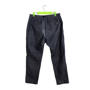 Op de afbeelding: Donkergrijze broek met een elastische tailleband en een trekkoord. De broek heeft twee achterzakken en een klein logo bij de rechterzak. De broek hangt aan een groen-witte hanger.