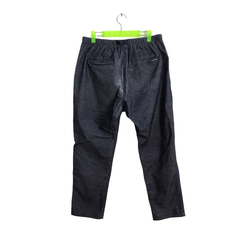 Op de afbeelding: Donkergrijze broek met een elastische tailleband en een trekkoord. De broek heeft twee achterzakken en een klein logo bij de rechterzak. De broek hangt aan een groen-witte hanger.