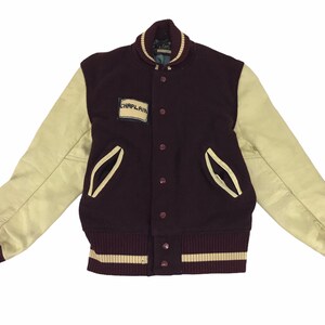 Puede incluir: Una chaqueta de béisbol granate con mangas de cuero crema y cuello y puños a rayas crema y granate. La chaqueta tiene un parche en el pecho izquierdo que dice "Chaplain".