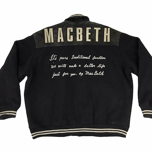 May include: Black varsity jacket with white and black striped trim. The back of the jacket has the word "MACBETH" in white lettering on a black suede patch. Below the word "MACBETH" is white text that reads "It's pure traditional fashion we will make a better life just for you. by Mac Buth."