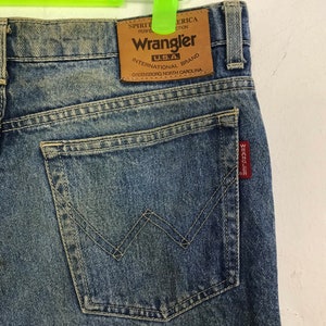 May include: Close-up of blue denim Wrangler jeans. The jeans feature a brown leather patch with the Wrangler logo and text. The back pocket has a signature W-shaped stitching. A red tag with white text is visible on the pocket.