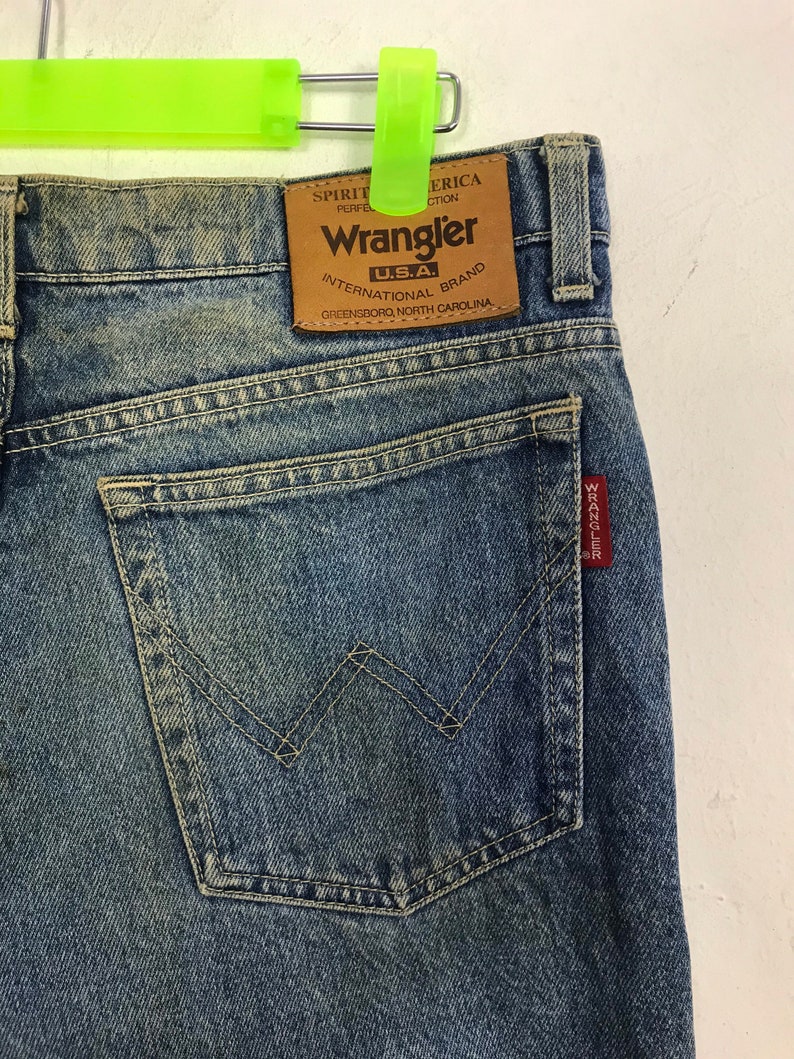 May include: Close-up of blue denim Wrangler jeans. The jeans feature a brown leather patch with the Wrangler logo and text. The back pocket has a signature W-shaped stitching. A red tag with white text is visible on the pocket.