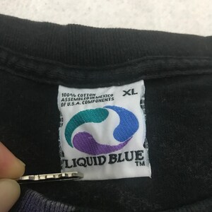 May include: Close-up of a black XL t-shirt tag. The tag is white with the Liquid Blue logo in teal, purple, and blue. The tag also includes the text "100% Cotton Assembled in Mexico of U.S.A. Components."