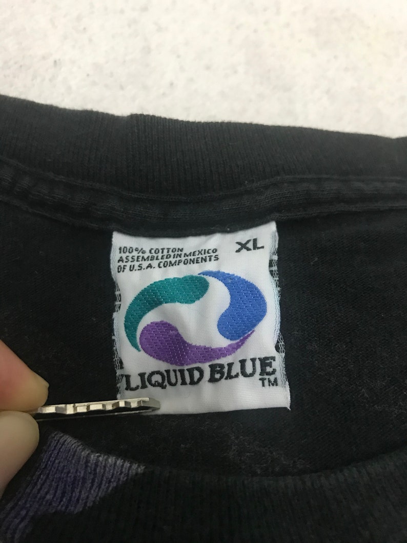 May include: Close-up of a black XL t-shirt tag. The tag is white with the Liquid Blue logo in teal, purple, and blue. The tag also includes the text "100% Cotton Assembled in Mexico of U.S.A. Components."