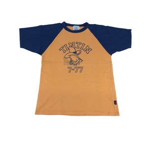 May include: A tan and navy blue raglan t-shirt featuring the cartoon character Tintin wearing a cowboy hat. The word "TINTIN" is arched above the character, with "7-77" printed below. The shirt has short sleeves.