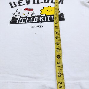Puede incluir: Camiseta blanca con el texto "DEVILOCK" sobre un dibujo de Hello Kitty y un sol amarillo. El texto "HELLO KITTY" est&aacute; debajo. La camiseta mide 61 cm de largo.
