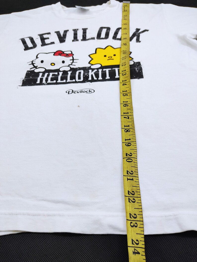 Puede incluir: Camiseta blanca con el texto "DEVILOCK" sobre un dibujo de Hello Kitty y un sol amarillo. El texto "HELLO KITTY" est&aacute; debajo. La camiseta mide 61 cm de largo.