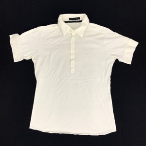 Op de afbeelding: Een wit shirt met korte mouwen, een kraag en een knoopsluiting. Het shirt heeft opgerolde mouwen en de tekst "A.A.P." is zichtbaar op de kraag.