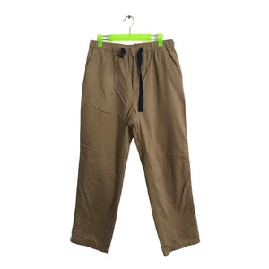 Può includere: Pantaloni color kaki con coulisse e chiusura con fibbia nera. I pantaloni sono appesi a una gruccia verde su sfondo bianco. I pantaloni hanno due tasche anteriori e una vestibilità ampia.