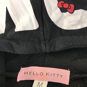 May include: Black hooded sweatshirt featuring a large white "Hello Kitty" graphic. The hood displays the character's face and bow. The pink label at the bottom reads "Hello Kitty" and indicates a size "M".
