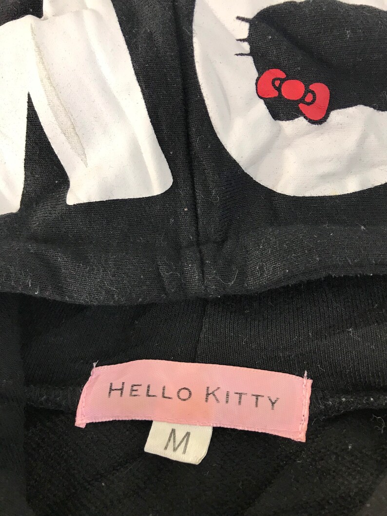 May include: Black hooded sweatshirt featuring a large white "Hello Kitty" graphic. The hood displays the character's face and bow. The pink label at the bottom reads "Hello Kitty" and indicates a size "M".