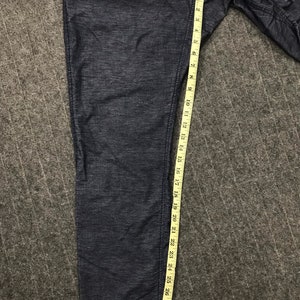 Op de afbeelding: Donkerblauwe spijkerbroek met een meetlint dat een lengte van ongeveer 76 cm aangeeft. De broek heeft een getextureerd uiterlijk en een klein label bij de tailleband. De broek is opgevouwen en ligt op een grijze ondergrond.