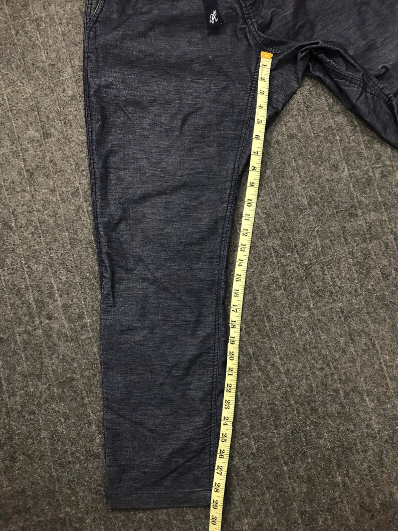 Op de afbeelding: Donkerblauwe spijkerbroek met een meetlint dat een lengte van ongeveer 76 cm aangeeft. De broek heeft een getextureerd uiterlijk en een klein label bij de tailleband. De broek is opgevouwen en ligt op een grijze ondergrond.