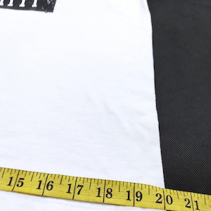 Puede incluir: Camiseta blanca con un gr&aacute;fico de un sol amarillo con una cara triste asom&aacute;ndose por encima de un rect&aacute;ngulo negro. El texto sobre el sol es parcialmente visible. Una cinta m&eacute;trica amarilla muestra el ancho de la camiseta, de 38 a 53 cm.