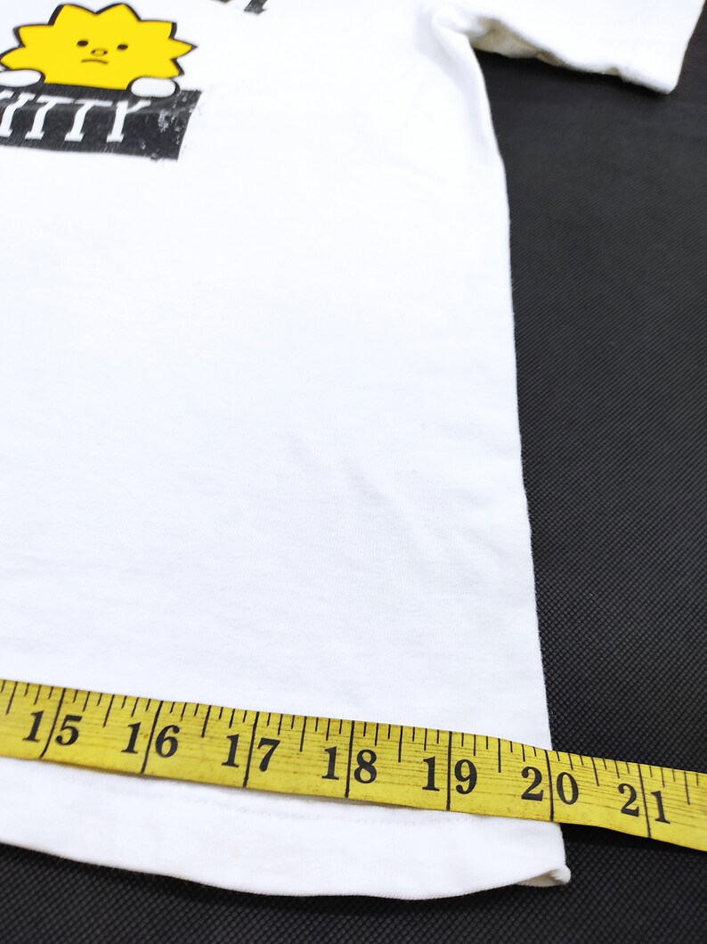 Puede incluir: Camiseta blanca con un gr&aacute;fico de un sol amarillo con una cara triste asom&aacute;ndose por encima de un rect&aacute;ngulo negro. El texto sobre el sol es parcialmente visible. Una cinta m&eacute;trica amarilla muestra el ancho de la camiseta, de 38 a 53 cm.