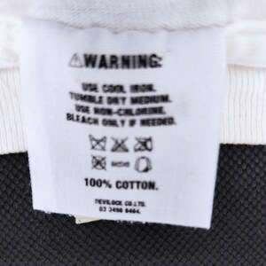 Puede incluir: Una etiqueta de ropa blanca con texto negro, que incluye la palabra "WARNING" en la parte superior. La etiqueta proporciona instrucciones de cuidado: usar plancha fr&iacute;a, secar en secadora a temperatura media, usar lej&iacute;a sin cloro solo si es necesario. La etiqueta es 100% algod&oacute;n.