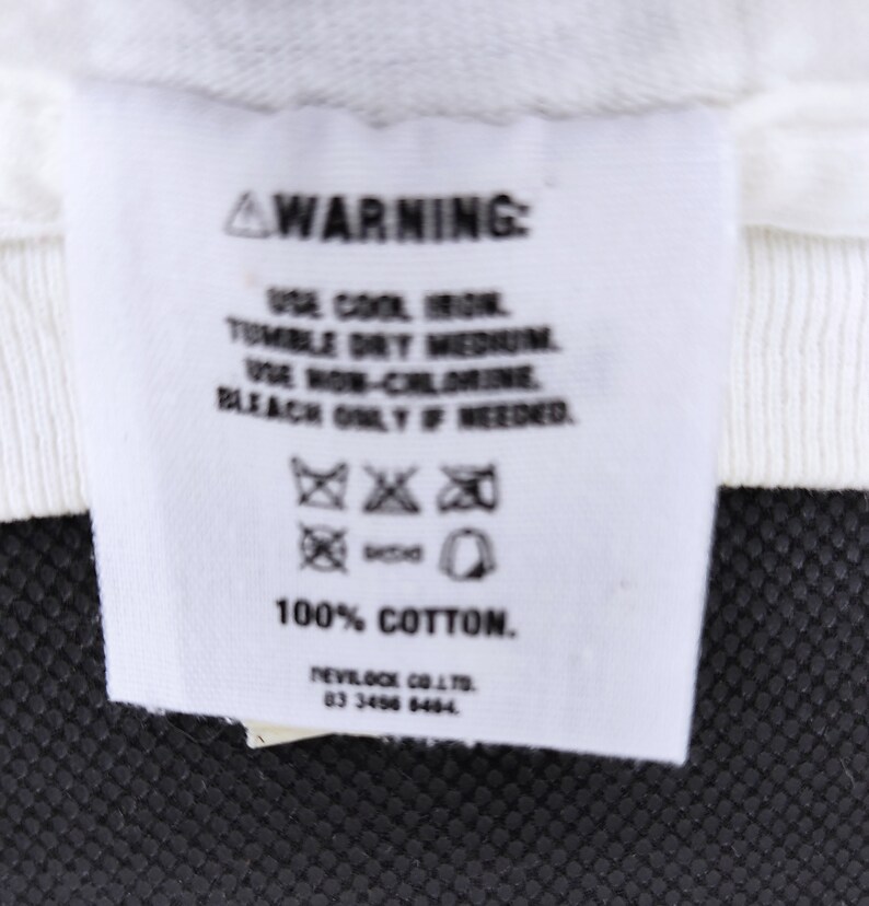 Puede incluir: Una etiqueta de ropa blanca con texto negro, que incluye la palabra "WARNING" en la parte superior. La etiqueta proporciona instrucciones de cuidado: usar plancha fr&iacute;a, secar en secadora a temperatura media, usar lej&iacute;a sin cloro solo si es necesario. La etiqueta es 100% algod&oacute;n.