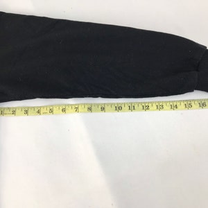 May include: Black long-sleeved shirt sleeve with a ribbed cuff. A yellow measuring tape is laid across the sleeve, showing measurements in inches. The sleeve is approximately 17 inches long.