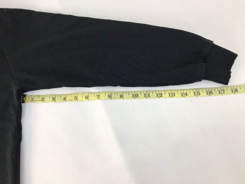 May include: Black long-sleeved shirt sleeve with a ribbed cuff. A yellow measuring tape is laid across the sleeve, showing measurements in inches. The sleeve is approximately 17 inches long.