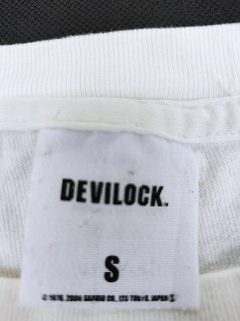 Puede incluir: Primer plano de una etiqueta de camiseta blanca con el nombre de la marca "DEVILOCK" en letras negras en negrita. Debajo del nombre de la marca est&aacute; la letra "S" en negro. La parte inferior de la etiqueta tiene texto que es dif&iacute;cil de leer.