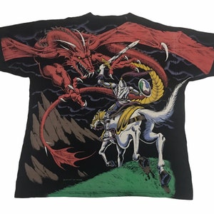 May include: Black t-shirt featuring a fantasy scene of a knight on horseback battling a red dragon. The knight is clad in silver armor and holds a sword. The dragon has large wings and sharp claws. The design is set against a dark background.