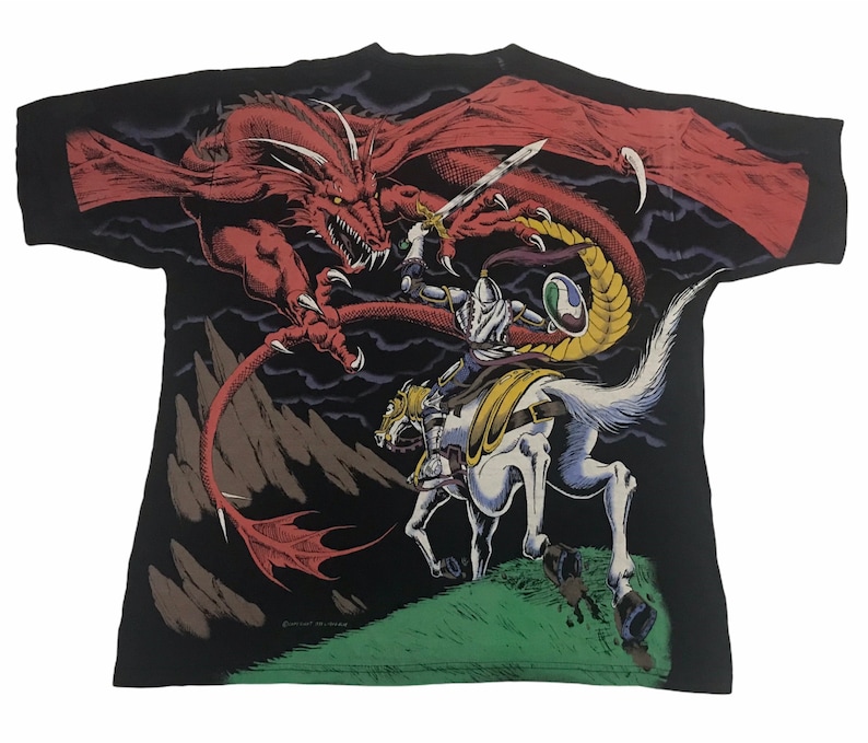 May include: Black t-shirt featuring a fantasy scene of a knight on horseback battling a red dragon. The knight is clad in silver armor and holds a sword. The dragon has large wings and sharp claws. The design is set against a dark background.