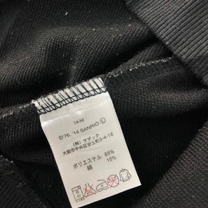 May include: Close-up of a black fabric with a white tag. The tag has Japanese text, the Sanrio copyright symbol, and washing instructions. The fabric is a blend of polyester and cotton.