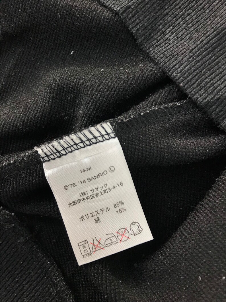 May include: Close-up of a black fabric with a white tag. The tag has Japanese text, the Sanrio copyright symbol, and washing instructions. The fabric is a blend of polyester and cotton.