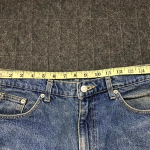 May include: Close-up of blue denim jeans with a button closure and pockets. A yellow measuring tape shows the waist size is approximately 40.6 cm. The jeans are on a grey surface.