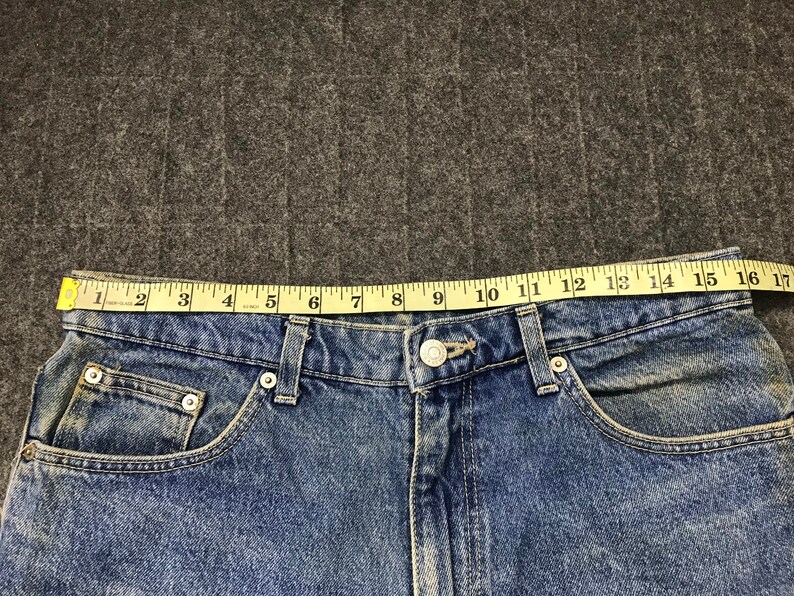 May include: Close-up of blue denim jeans with a button closure and pockets. A yellow measuring tape shows the waist size is approximately 40.6 cm. The jeans are on a grey surface.