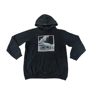 Op de afbeelding: Zwarte hoodie met een voorzak en een grafische print. De print is een zwart-wit afbeelding van een stadsgezicht met een cirkelvormig ontwerp en de tekst "@" en "EE.UU. 2000".