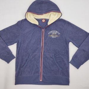 Puede incluir: Sudadera azul marino con cremallera y capucha forrada de forro polar color crema. El logotipo y el texto de Converse están bordados en el pecho. La cremallera tiene rayas rojas y blancas. Una prenda de abrigo clásica y cómoda.