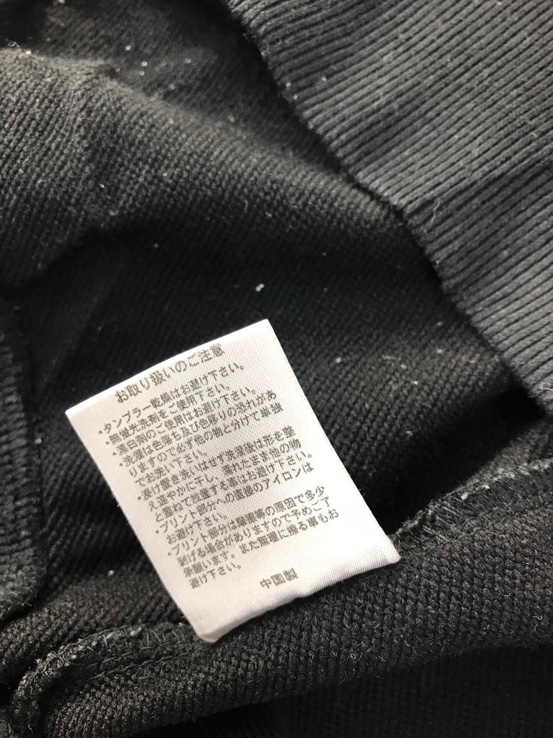 May include: Close-up of a black fabric with a white tag attached. The tag has Japanese text and care instructions. The fabric appears to be a dark, textured material, possibly a sweatshirt or similar garment.