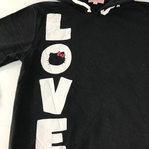 May include: Black hooded sweatshirt featuring the word "LOVE" in large white letters down the front. The letter "O" has a Hello Kitty graphic. The hood has the word "LOVE" printed on it. The sweatshirt has a drawstring and a pink tag.