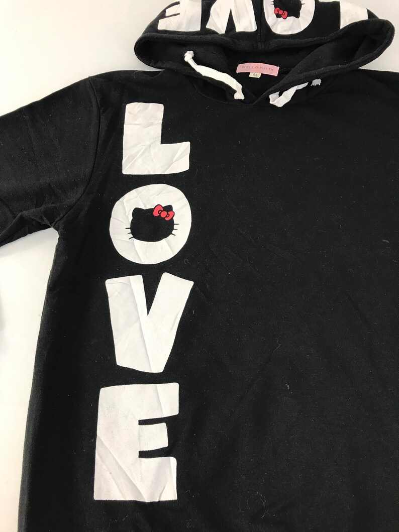 May include: Black hooded sweatshirt featuring the word "LOVE" in large white letters down the front. The letter "O" has a Hello Kitty graphic. The hood has the word "LOVE" printed on it. The sweatshirt has a drawstring and a pink tag.