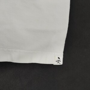 Puede incluir: Primer plano de una camiseta blanca. El dobladillo inferior es visible, con una peque&ntilde;a etiqueta con un logotipo de calavera y huesos cruzados. La tela parece suave y lisa, con una textura sutil.