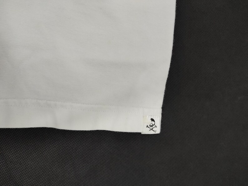 Puede incluir: Primer plano de una camiseta blanca. El dobladillo inferior es visible, con una peque&ntilde;a etiqueta con un logotipo de calavera y huesos cruzados. La tela parece suave y lisa, con una textura sutil.