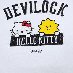 Puede incluir: Camiseta blanca con la inscripci&oacute;n "DEVILOCK" sobre un dibujo de Hello Kitty con un lazo rojo y un personaje amarillo. Debajo, el texto "HELLO KITTY" est&aacute; impreso en un rect&aacute;ngulo negro. El logotipo de Devilock est&aacute; impreso debajo.