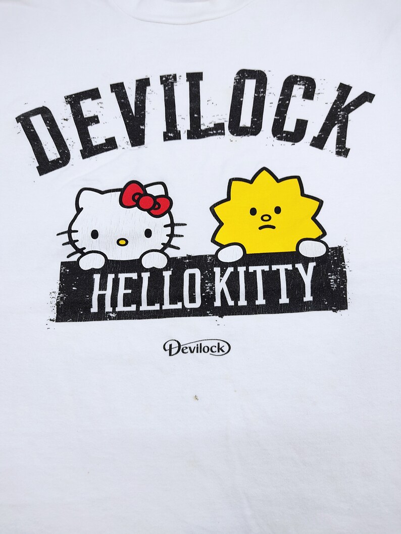 Puede incluir: Camiseta blanca con la inscripci&oacute;n "DEVILOCK" sobre un dibujo de Hello Kitty con un lazo rojo y un personaje amarillo. Debajo, el texto "HELLO KITTY" est&aacute; impreso en un rect&aacute;ngulo negro. El logotipo de Devilock est&aacute; impreso debajo.