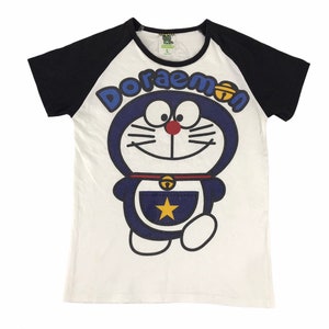 May include: White and black raglan sleeve t-shirt featuring a large graphic of the cartoon character Doraemon. The blue Doraemon has a red nose, a bell collar, and a yellow star on its pocket. The word "Doraemon" is written in blue letters.