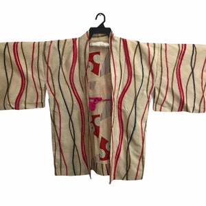 May include: A beige kimono with a wavy, abstract pattern in red, black, and brown. The kimono has a pink tassel hanging from the front.