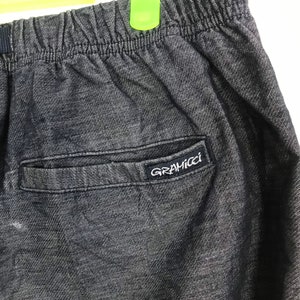 Op de afbeelding: Close-up van een donkergrijze Gramicci broek. De broek heeft een elastische tailleband en een achterzak met het Gramicci-logo in het wit. De stof lijkt een getextureerde denim te zijn.