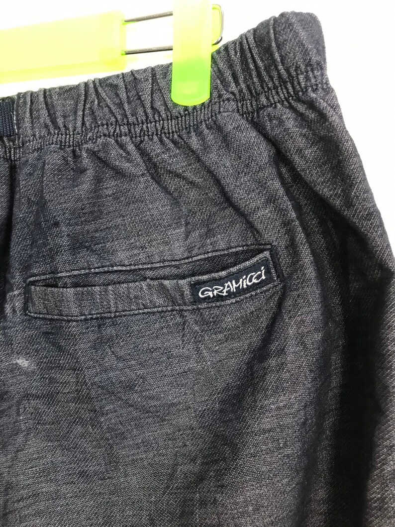 Op de afbeelding: Close-up van een donkergrijze Gramicci broek. De broek heeft een elastische tailleband en een achterzak met het Gramicci-logo in het wit. De stof lijkt een getextureerde denim te zijn.