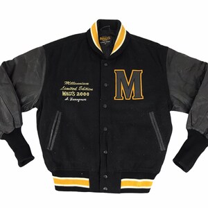 Puede incluir: Chaqueta de béisbol negra con mangas de cuero. La chaqueta tiene un ribete de rayas doradas y blancas alrededor del cuello, los puños y el dobladillo. La parte delantera de la chaqueta presenta una gran "M" bordada en oro y el texto "Millennium Limited Edition MAITS 2000 S. Yamaguti".
