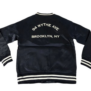 May include: A navy blue satin bomber jacket with white trim on the collar, cuffs, and hem. The back of the jacket features the text "94 Wythe Ave Brooklyn, NY" in white lettering. The jacket has long sleeves.