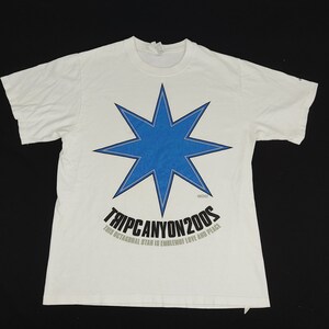 Puede incluir: Camiseta blanca con un gran diseño de estrella azul de ocho puntas y un contorno azul. Debajo de la estrella está el texto "TRIP CANYON 2005" y "THIS OCTAGONAL STAR IS EMBLEM OF LOVE AND PEACE".