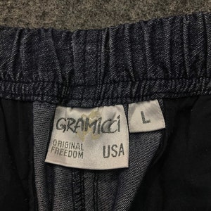 Op de afbeelding: Close-up van een donkerblauwe spijkerbroek. De tailleband is elastisch. Een wit label toont de merknaam "Gramicci", de woorden "Original Freedom" en "USA". De maat "L" is ook zichtbaar.