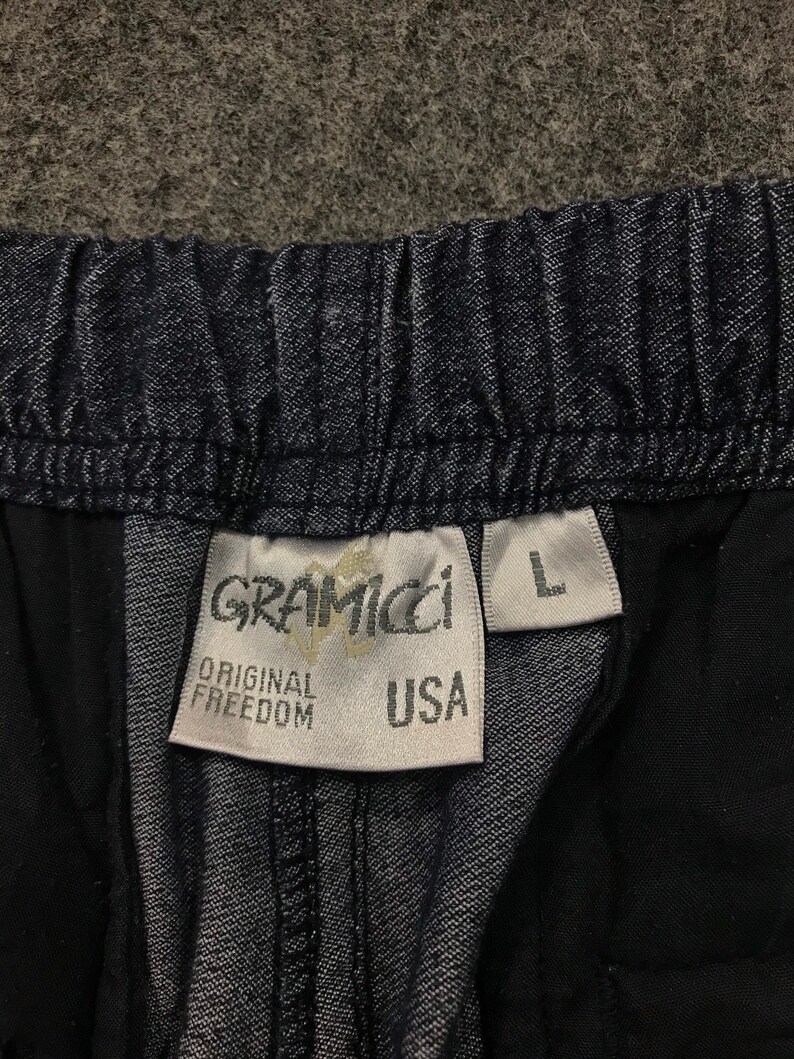 Op de afbeelding: Close-up van een donkerblauwe spijkerbroek. De tailleband is elastisch. Een wit label toont de merknaam "Gramicci", de woorden "Original Freedom" en "USA". De maat "L" is ook zichtbaar.