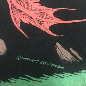 May include: Close-up of a black t-shirt featuring a red wing graphic. The text "COPYRIGHT 1993 LIQUID BLUE" is printed in green. The design is set against a green and gray background.