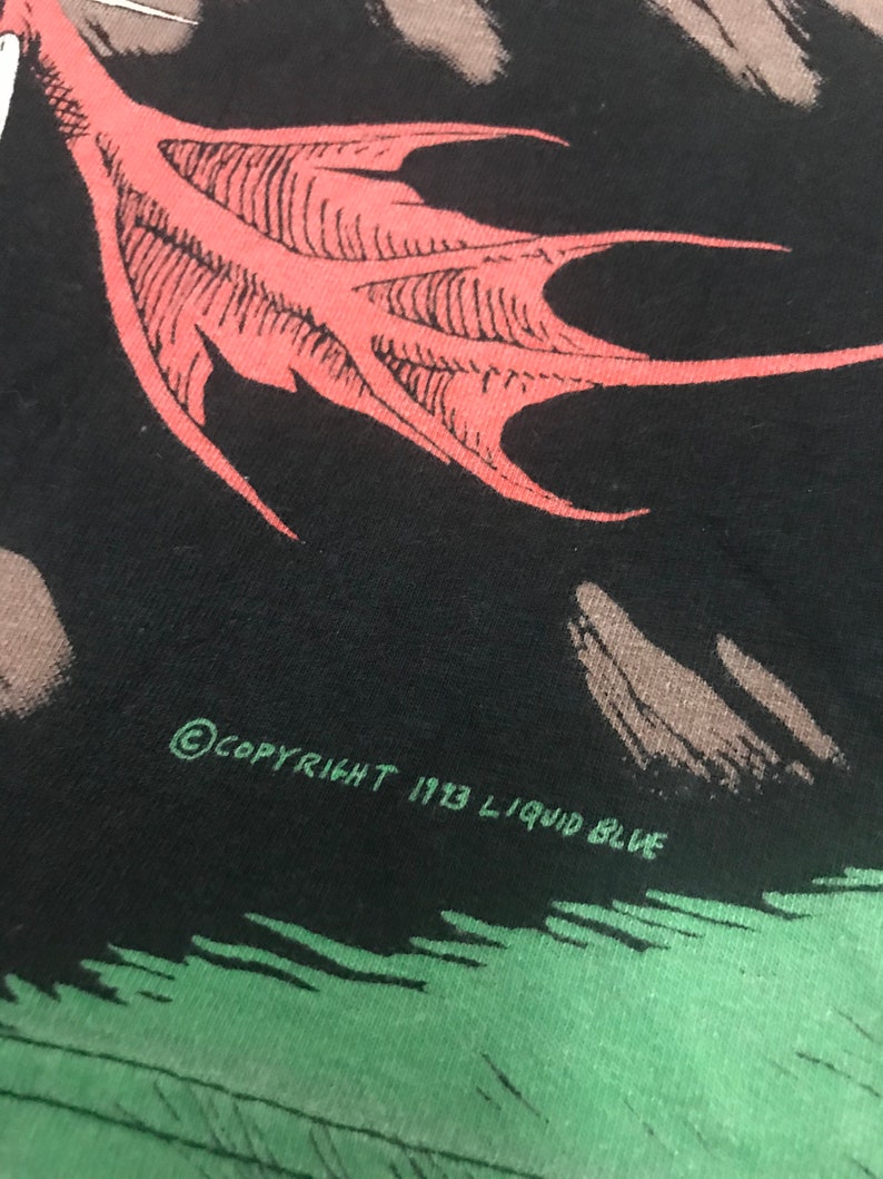 May include: Close-up of a black t-shirt featuring a red wing graphic. The text "COPYRIGHT 1993 LIQUID BLUE" is printed in green. The design is set against a green and gray background.